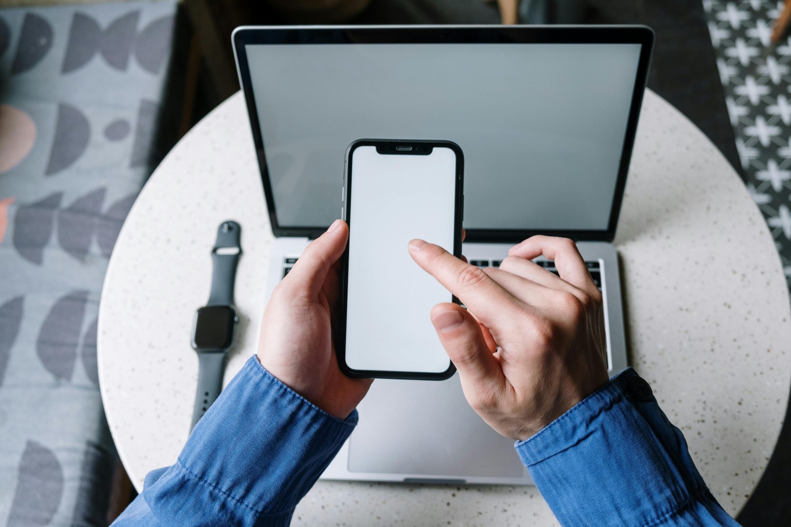 Hands operating a smartphone above a laptop with a smartwatch nearby, showcasing modern technology.